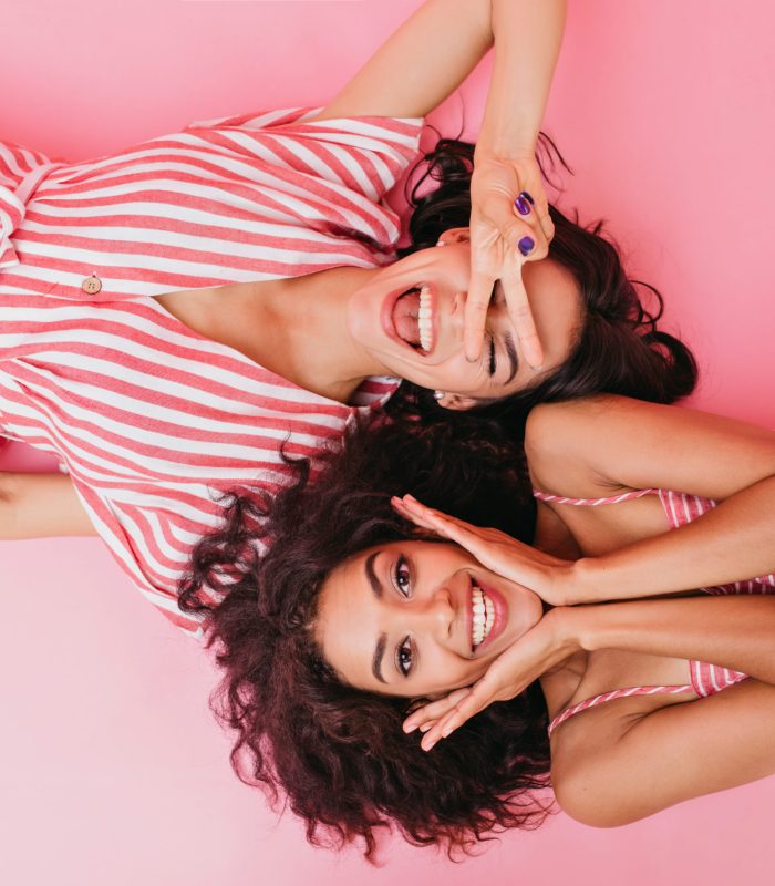 On pink isolated background beautiful dark-skinned girls with cute smiles are having fun and posing in front of camera lying on their backs